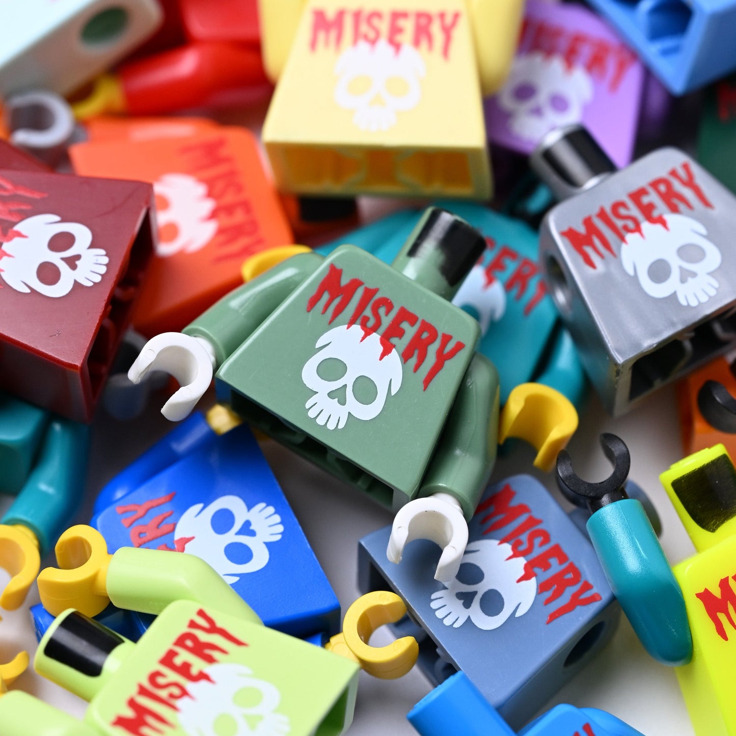 A pile of colorful toy minifigure torso pieces printed with the word 'MISERY' and a white skull graphic, shown in multiple colors including blue, green, yellow, red, gray, and purple.