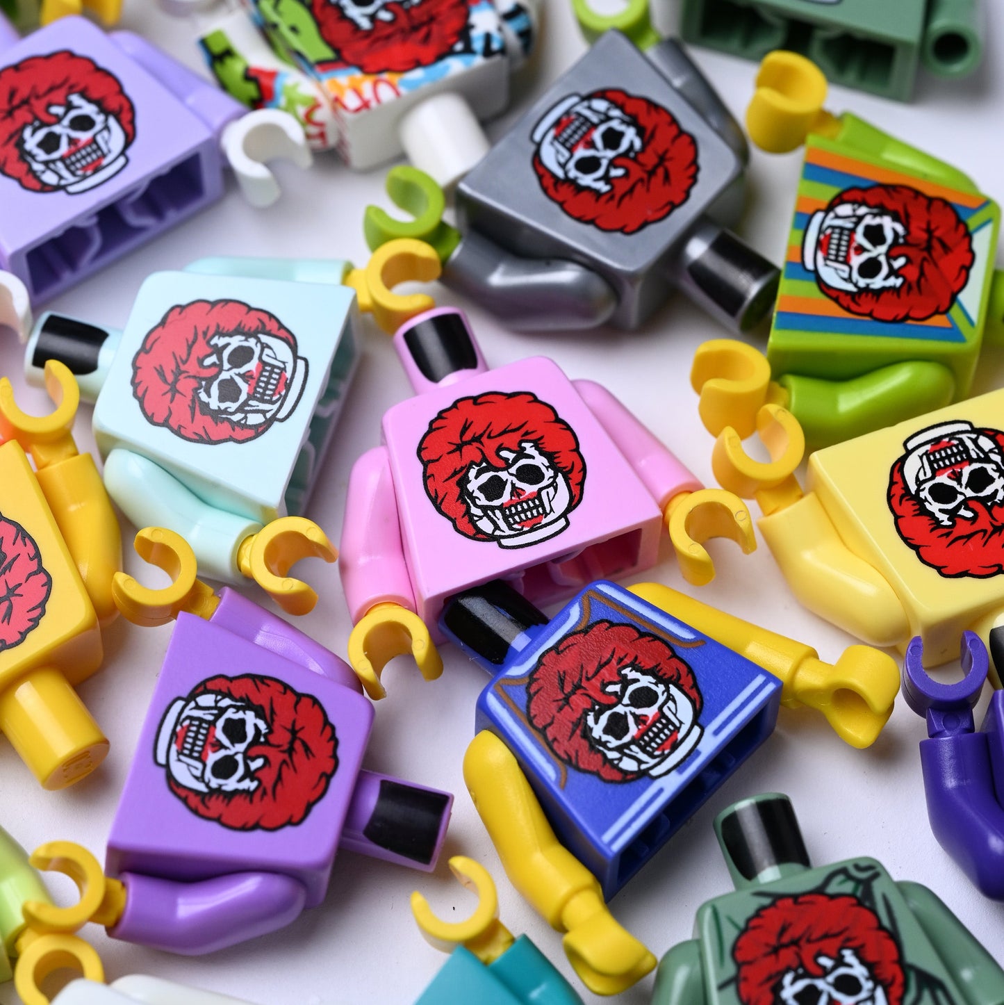 A close-up, top-down view of multiple colorful toy minifigures with printed skull graphics on their chests, scattered across a white background. Kind of like a creepy ronald mcdonald zombie.