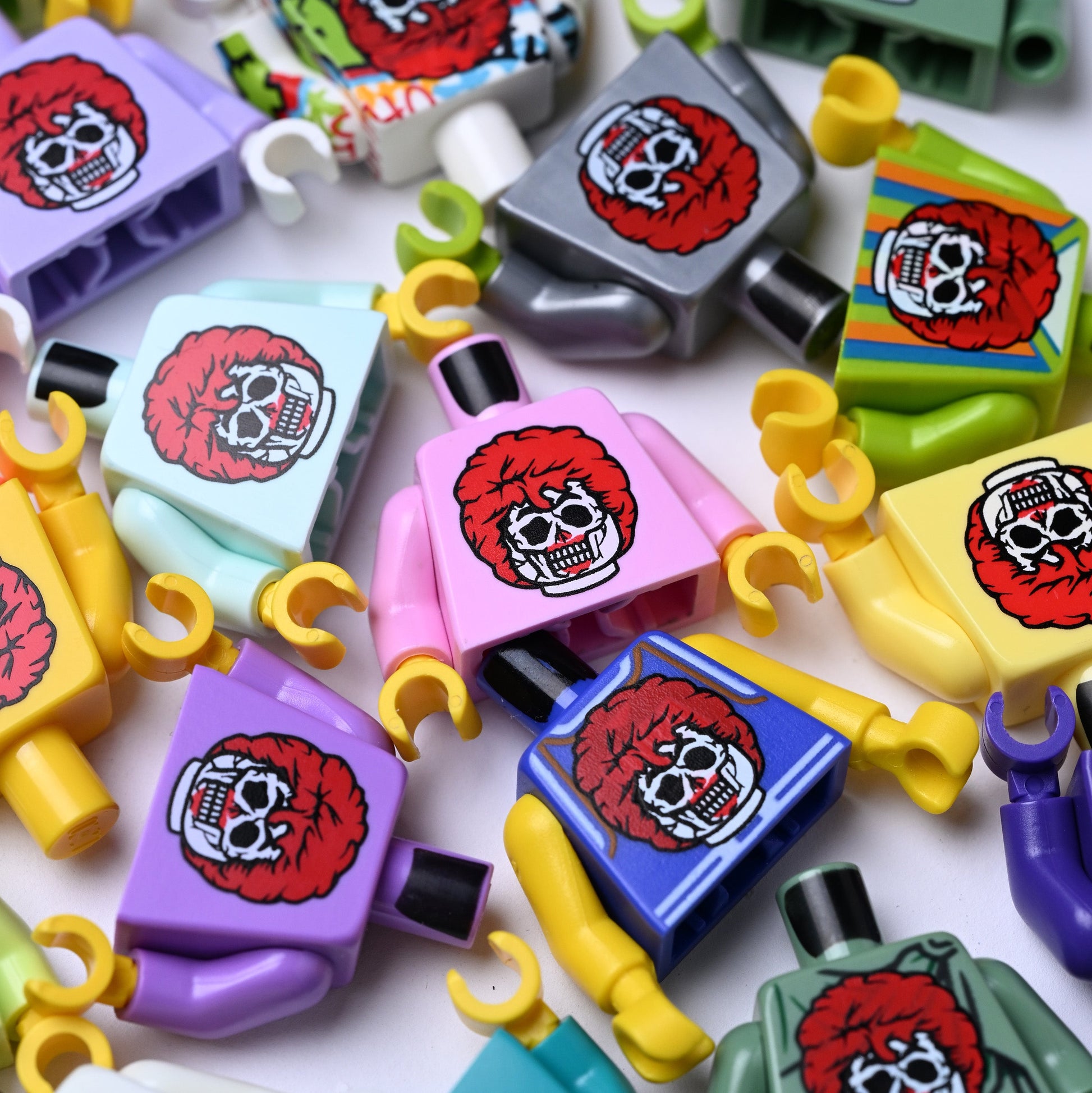A close-up, top-down view of multiple colorful toy minifigures with printed skull graphics on their chests, scattered across a white background. Kind of like a creepy ronald mcdonald zombie.