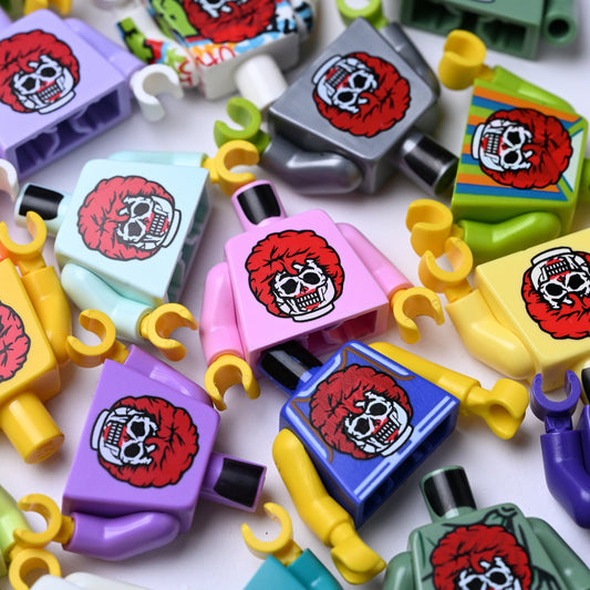 A close-up, top-down view of multiple colorful toy minifigures with printed skull graphics on their chests, scattered across a white background. Kind of like a creepy ronald mcdonald zombie.